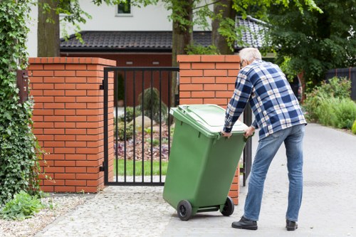 A man with grey hair, wearing a blue and white checkered shirt and blue jeans, is pushing a large green plastic rubbish bin on wheels toward a black metal gate. The gate is situated between two red brick pillars, leading into a residential property. The bin appears to contain waste and is positioned on a paved driveway with a gravel path nearby. In the background, there are lush green trees and shrubs, and part of a modern house with white walls and window frames is visible behind the garden. The scene is well-lit with natural daylight, indicating a clear day, and the surroundings suggest a typical suburban setting in Camden Town, where commercial waste removal services by Commercial Waste Camden Town could be relevant for waste disposal solutions.