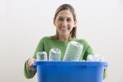 A young woman with shoulder-length light brown hair and a bright smile is holding a blue recycling bin filled with empty plastic bottles of various sizes and shapes, all made of clear transparent plastic with visible ridges and caps. She is wearing a green sweater and standing against a plain white background. The plastic bottles appear clean and are loosely arranged in the bin, which is positioned at chest height, making it easy for her to hold and display them prominently. The setting suggests an indoor environment, possibly a residential or office space. This image subtly highlights the importance of recycling waste such as plastic bottles, aligning with rubbish removal services offered by companies like Commercial Waste Camden Town, which serve the Camden Town area and surrounding postcode regions. The focus on plastic waste emphasizes environmentally conscious waste management practices and the importance of proper disposal of recyclable materials.