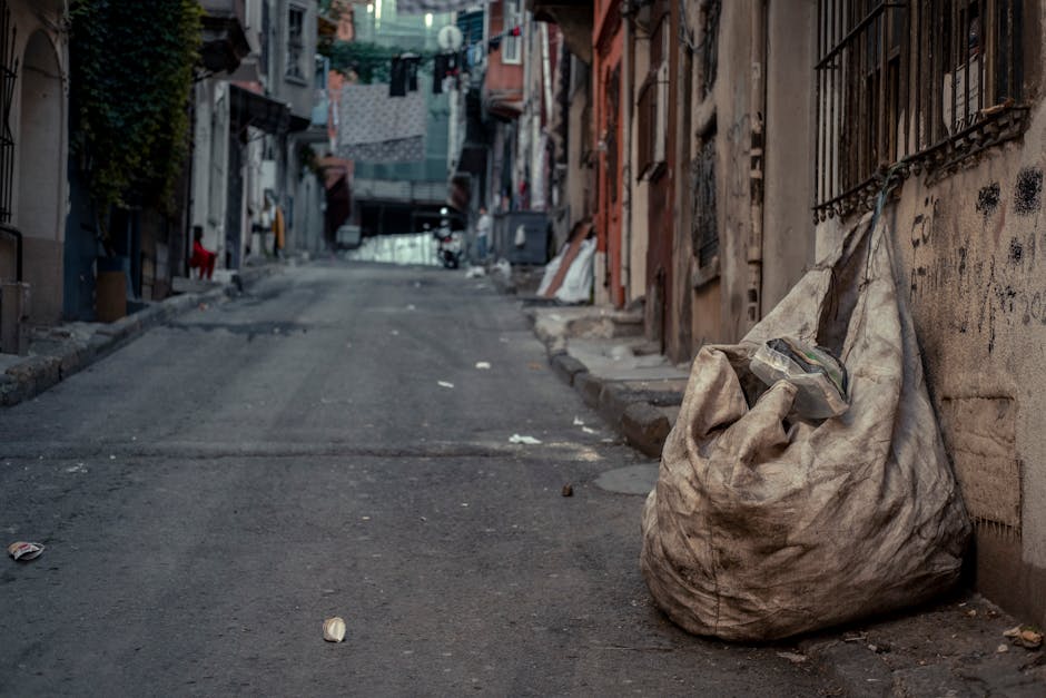 Two large black plastic bags of rubbish, heavily crumpled and tied at the top, are placed on the pavement near a black metal fence. The bags appear to contain household waste and are positioned close 