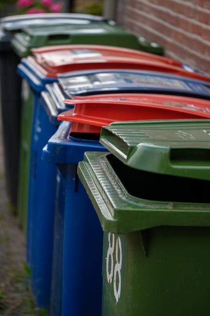 Close-up of a large black plastic rubbish bag tied at the top, placed against a plain light-colored wall on a paved surface, with other similar bags partly visible on the left. The bag appears to cont