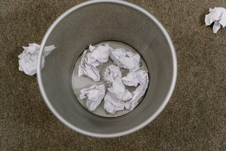 An overhead view of a cylindrical metal rubbish bin with a smooth, matte finish, placed on a brown textured carpet. Inside the bin, there are several crumpled white sheets of paper, some partially sub