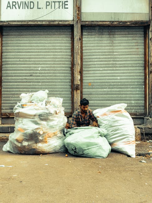 A high-angle view of a pile of mixed waste materials scattered across a ground surface, including large black and white garbage bags filled with rubbish, cardboard boxes, wooden spools, and various lo