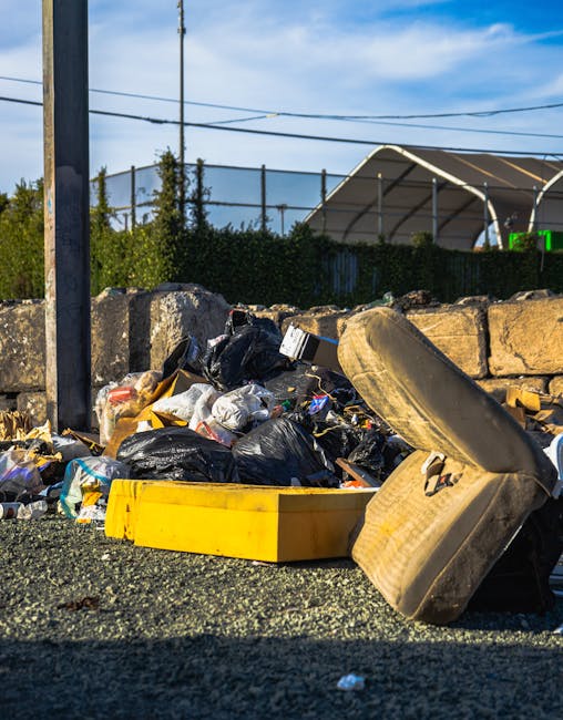 A close-up view of an outdoor heap of mixed rubbish materials accumulated against a low stone wall and gravel ground. Visible in the pile are black and white plastic bin bags, some torn open revealing